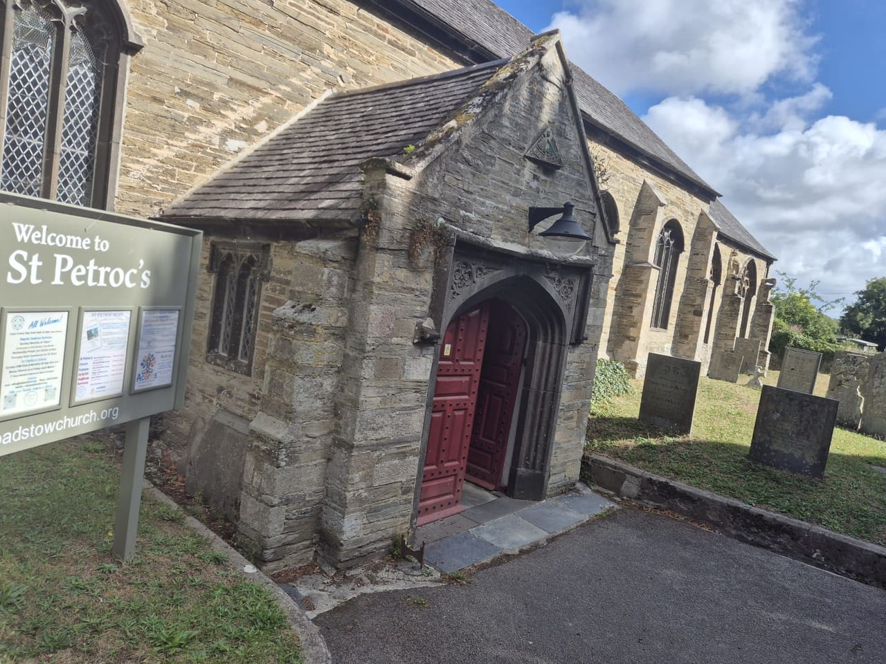 The arched stone porch and red door of St Petroc's Church, framed by ivy and gravestones